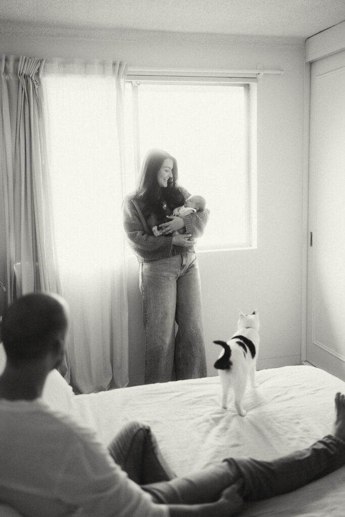 Parents holding baby Maisie in a sun filled bedroom while their cat watches. 